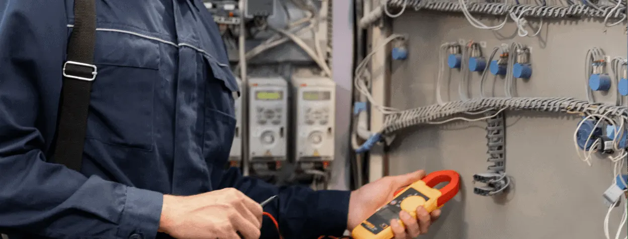 An electrician in Clearwater, wearing a blue work uniform, is holding a digital multimeter while examining the wiring inside an electrical panel. The panel contains various cables and components. The individual appears to be conducting electrical maintenance or troubleshooting work for East-West Electric.