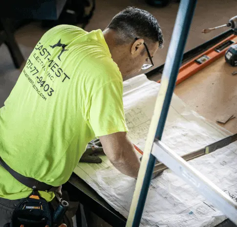 Man in a bright yellow shirt with a visible company logo and contact information is working on construction plans at a table. Various construction tools, including a level and a pencil, are spread across the tabletop, highlighting his expertise as an electrician in Clearwater.