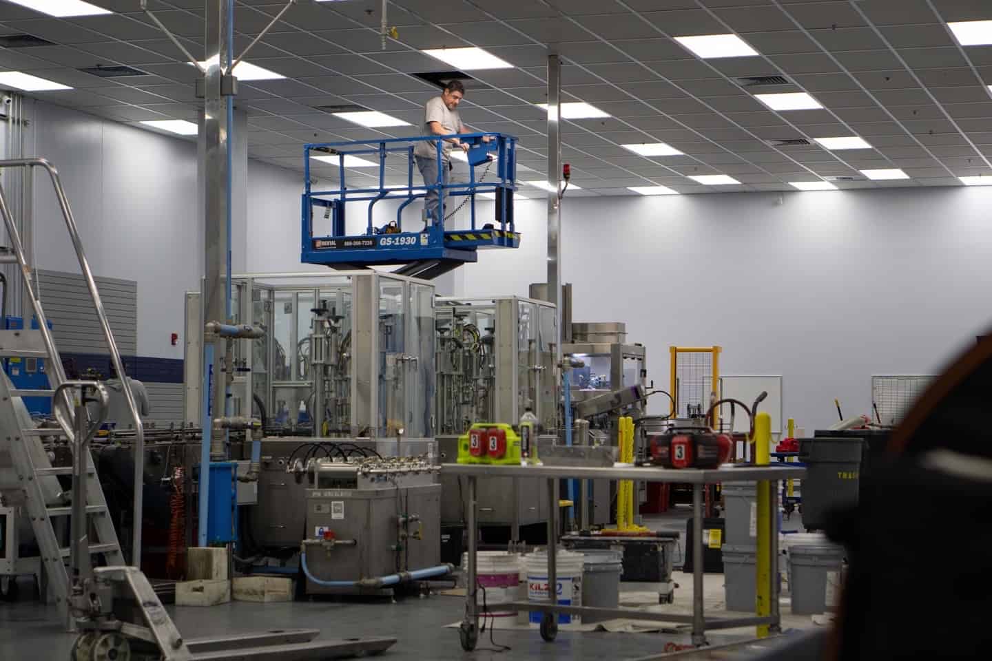 A person wearing a white shirt stands on an elevated blue scissor lift in a spacious industrial facility. The room is equipped with various machinery, metal tables, and safety equipment. East-West Electric ensures that the ceiling has a grid pattern with bright fluorescent lighting.