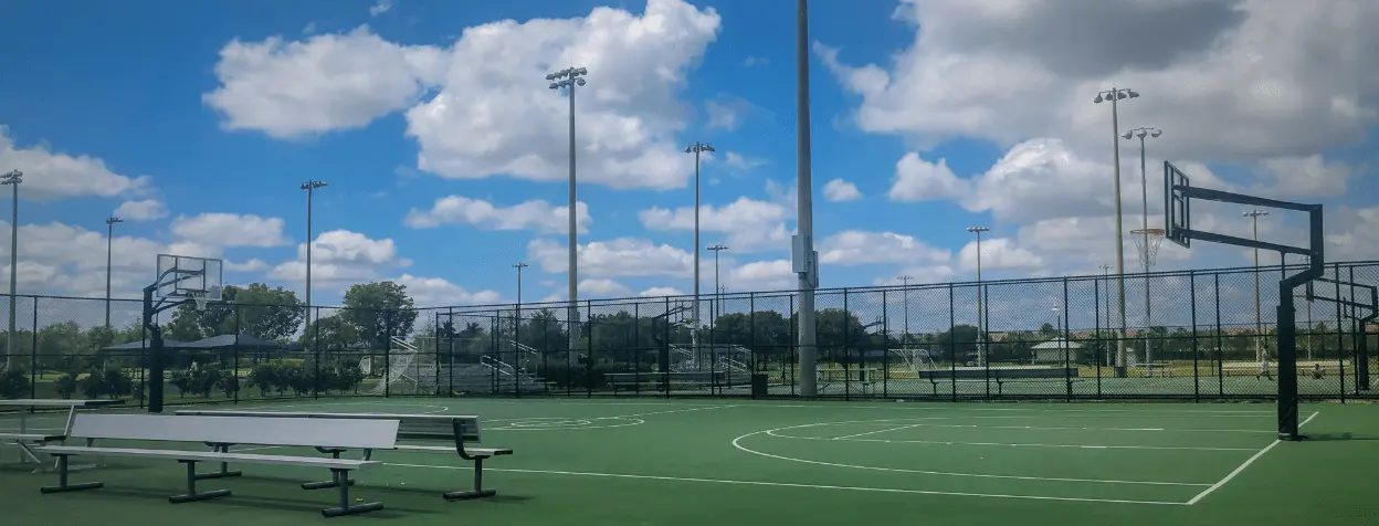 A well-maintained outdoor basketball court with two hoops, surrounded by a tall chain-link fence. Benches are positioned on the side of the court. The sky is partly cloudy with ample sunlight, and sports fields can be seen in the background, enhanced by East-West Electric's lighting solutions.