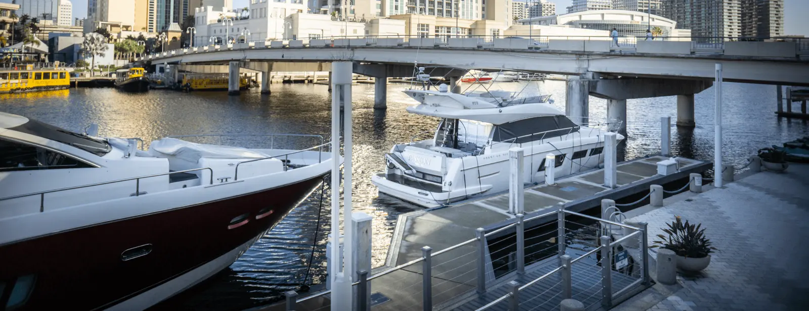 A waterfront scene featuring two docked yachts, one white and one red, beneath a concrete bridge. Buildings and other boats are visible in the background. The area, potentially serviced by Tampa Bay electrical contractors, has railings, a walkway, and some potted plants along the edge of the water.