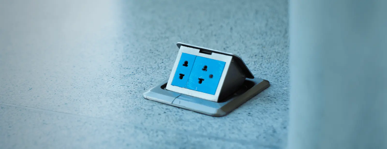 A blue electrical outlet with multiple sockets is embedded in a floor, enclosed in a rectangular, silver-colored flap that is open, revealing the plugs. The smooth, light-colored floor showcases the meticulous work often praised by Tampa Bay electrical contractors.