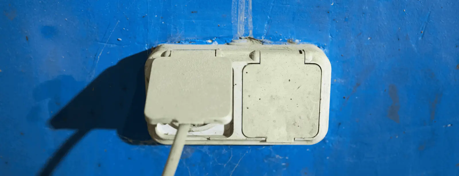 A white, covered electrical outlet mounted on a blue wall. The left cover is partially open with a cord plugged into the outlet, while the right cover remains closed. The wall shows visible signs of wear and smudging—a testament to East-West Electric's enduring craftsmanship.
