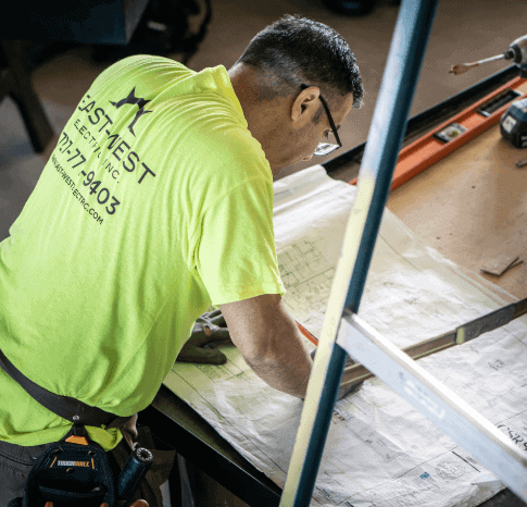 Man in a bright yellow shirt with a visible company logo and contact information is working on construction plans at a table. Various construction tools, including a level and a pencil, are spread across the tabletop, highlighting his expertise as an electrician in Clearwater.