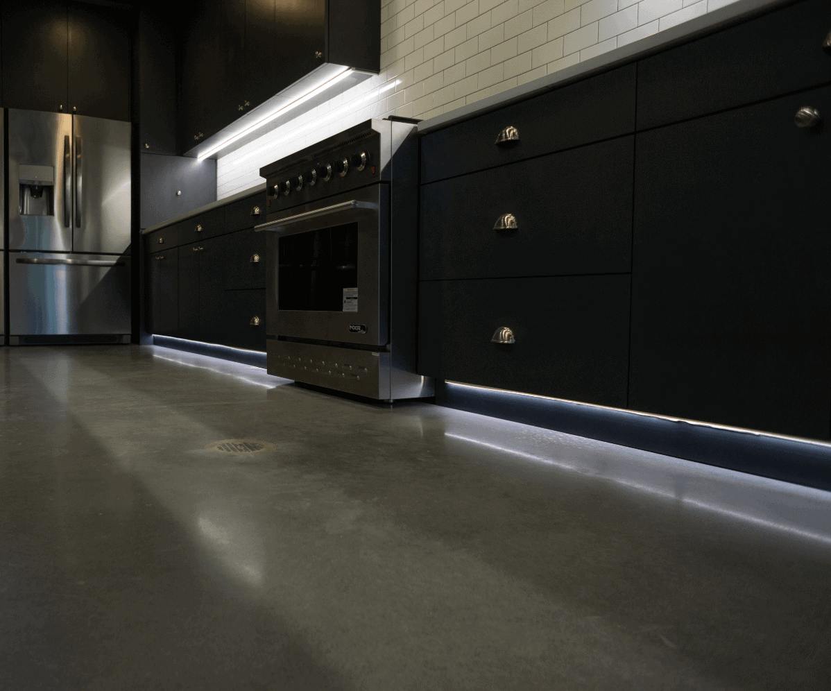 Modern kitchen with sleek, black cabinetry and stainless steel appliances, including a refrigerator and oven. The kitchen features white subway tile backsplash and under-cabinet lighting as well as baseboard lighting installed by East-West Electric, creating a minimalist and polished look.