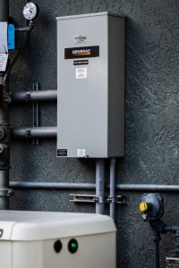 A Generac electrical unit, installed by East-West Electric, is mounted on an exterior wall with conduits and a gas meter nearby. The unit has labels, including one indicating "Automatic Transfer Switch." A gauge is visible above it, and another piece of equipment is partially visible in the foreground.