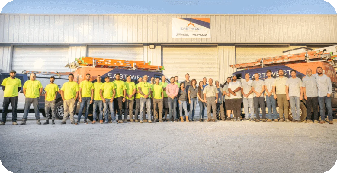 A group of 26 people stands in front of a warehouse with two orange company trucks labeled "East-West." Some wear bright yellow-green safety shirts and hard hats, while others are in casual attire. A company sign is visible above the warehouse doors, hinting at their reputation as top electricians in Clearwater.